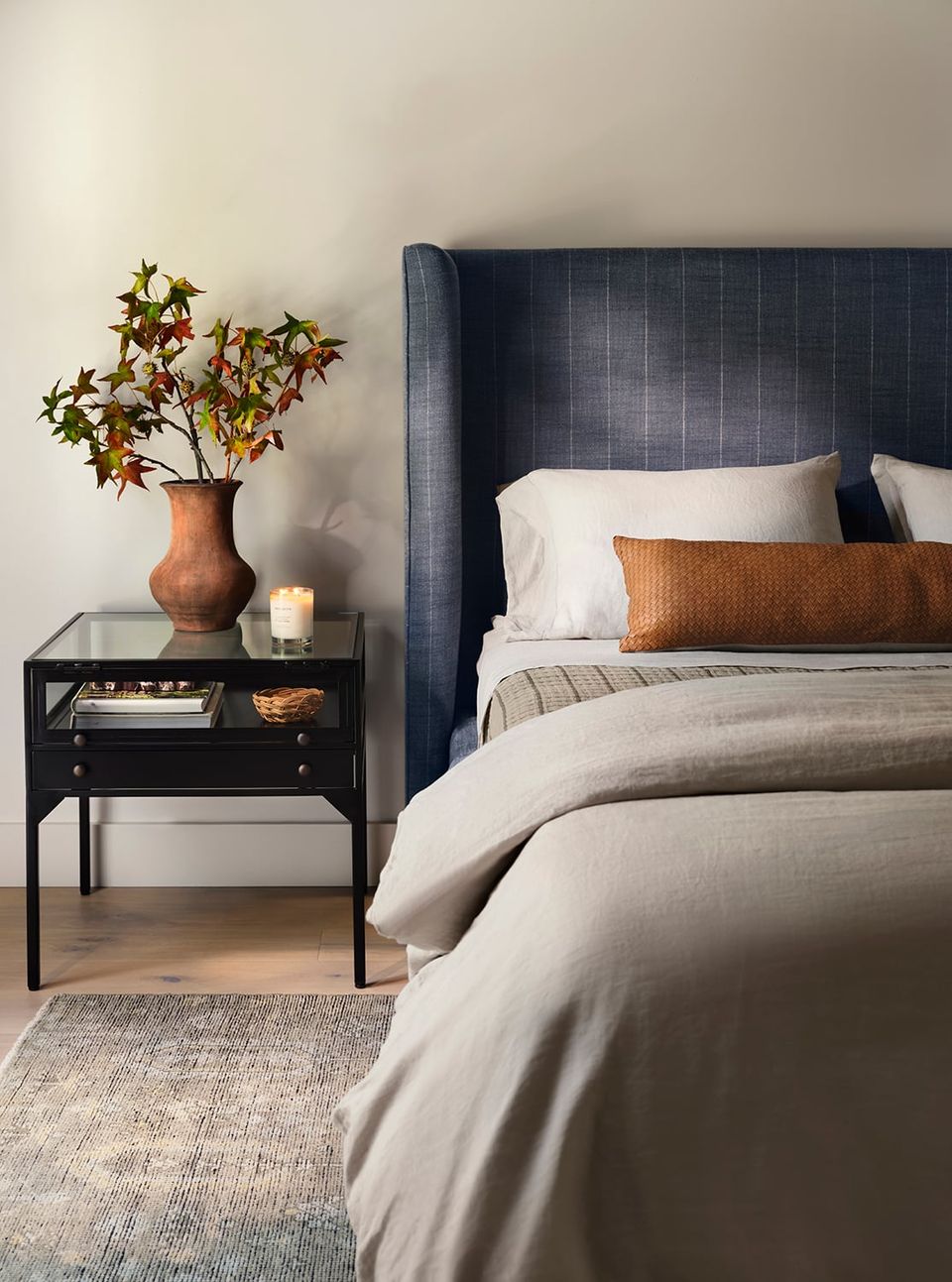 Contemporary bedroom with navy upholstered headboard, warm terracotta pillow accent, neutral layered bedding, and minimalist black nightstand