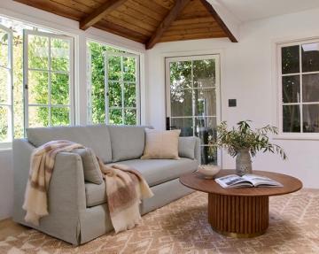 Bright sunlit living room with coastal farmhouse style: gray linen sofa, wood ceiling beams, round wood coffee table, and natural textiles.