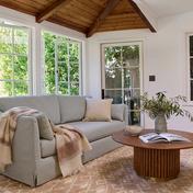 Bright sunlit living room with coastal farmhouse style: gray linen sofa, wood ceiling beams, round wood coffee table, and natural textiles.