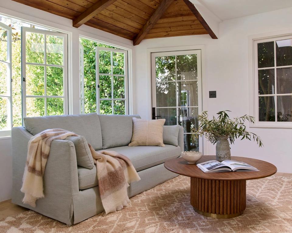 Bright organic modern space with gray upholstered sofa, wooden ceiling beams, ribbed wood coffee table, and potted greenery