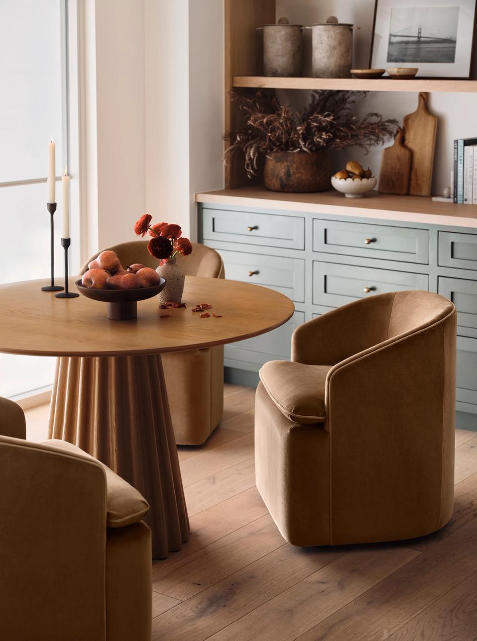 Warm minimalist dining space with natural wood round table, caramel leather chairs, and soft blue storage cabinet in earthy tones