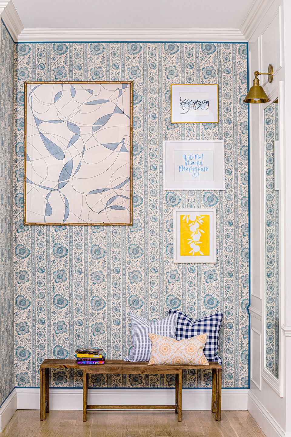 Entryway with blue and cream patterned wallpaper, gallery wall of framed botanical art, wooden bench with navy gingham and patterned pillows, brass sconce.