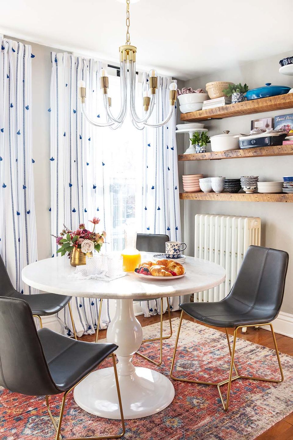 Mid-century dining nook with patterned curtains, round white table, black chairs, and open shelving combining style and storage.