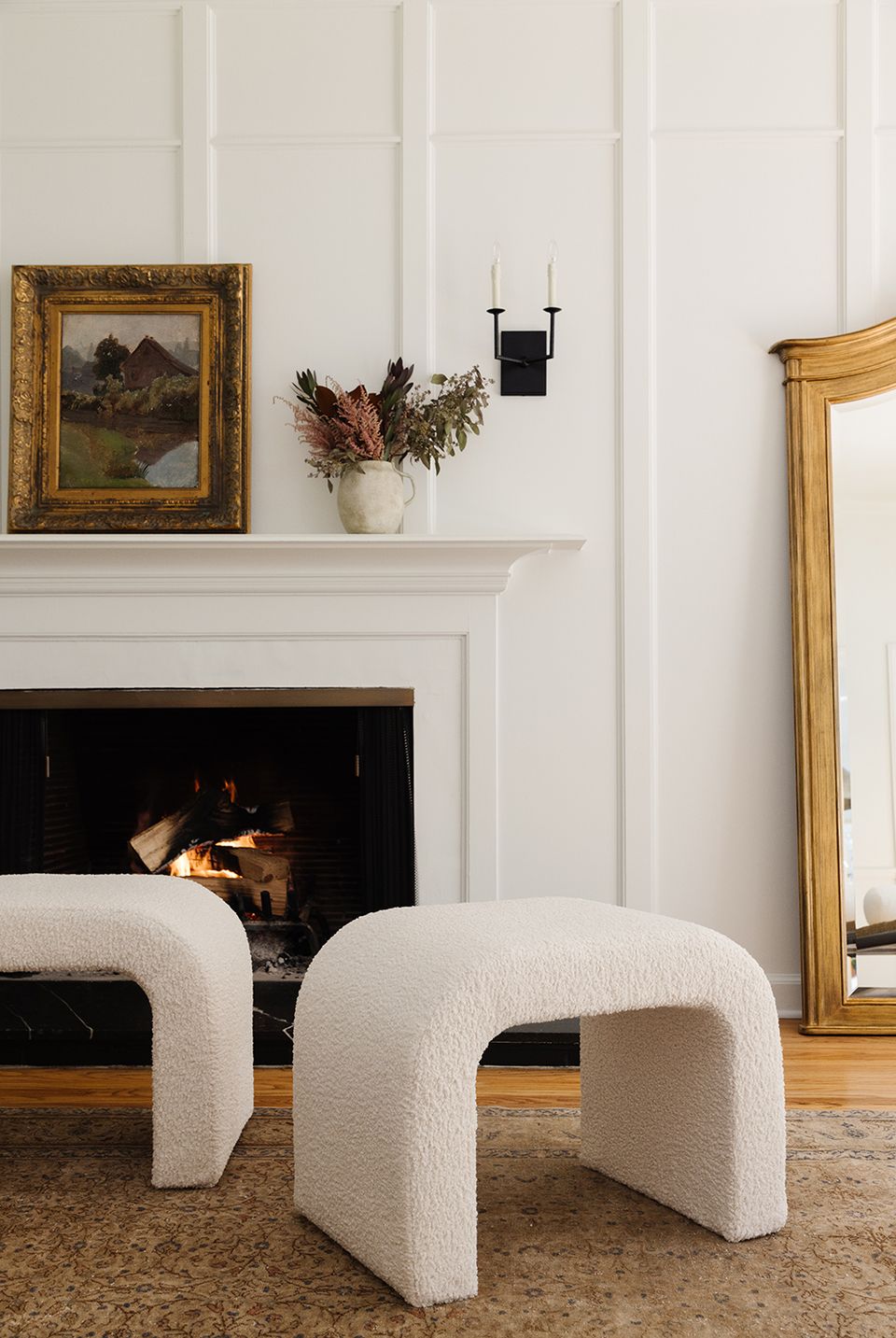 Contemporary fireplace mantel with ornate gold-framed landscape art, cream sculptural stool, and minimal black accents