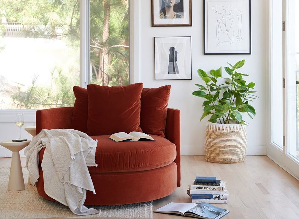 Cozy rust-red round chair corner with soft throws, layered artwork, and lush potted plant in bright alcove.