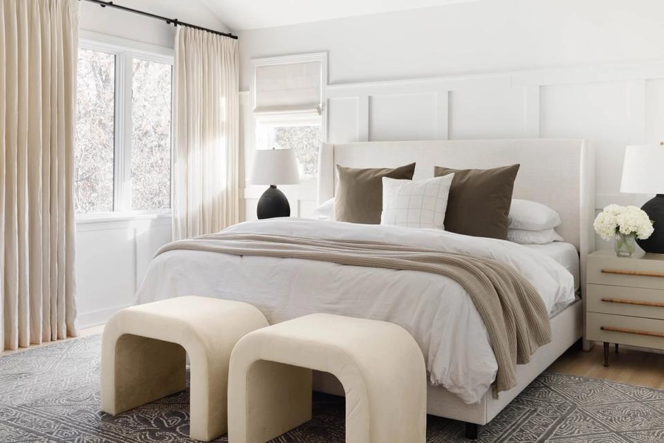 Contemporary organic modern bedroom with white paneled headboard, warm neutral palette, cream ottomans, and soft layered textures
