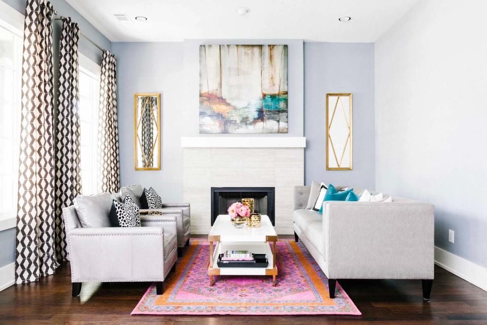 Bright transitional living room with soft blue walls, geometric patterned curtains, white seating, and jewel-toned accents around fireplace