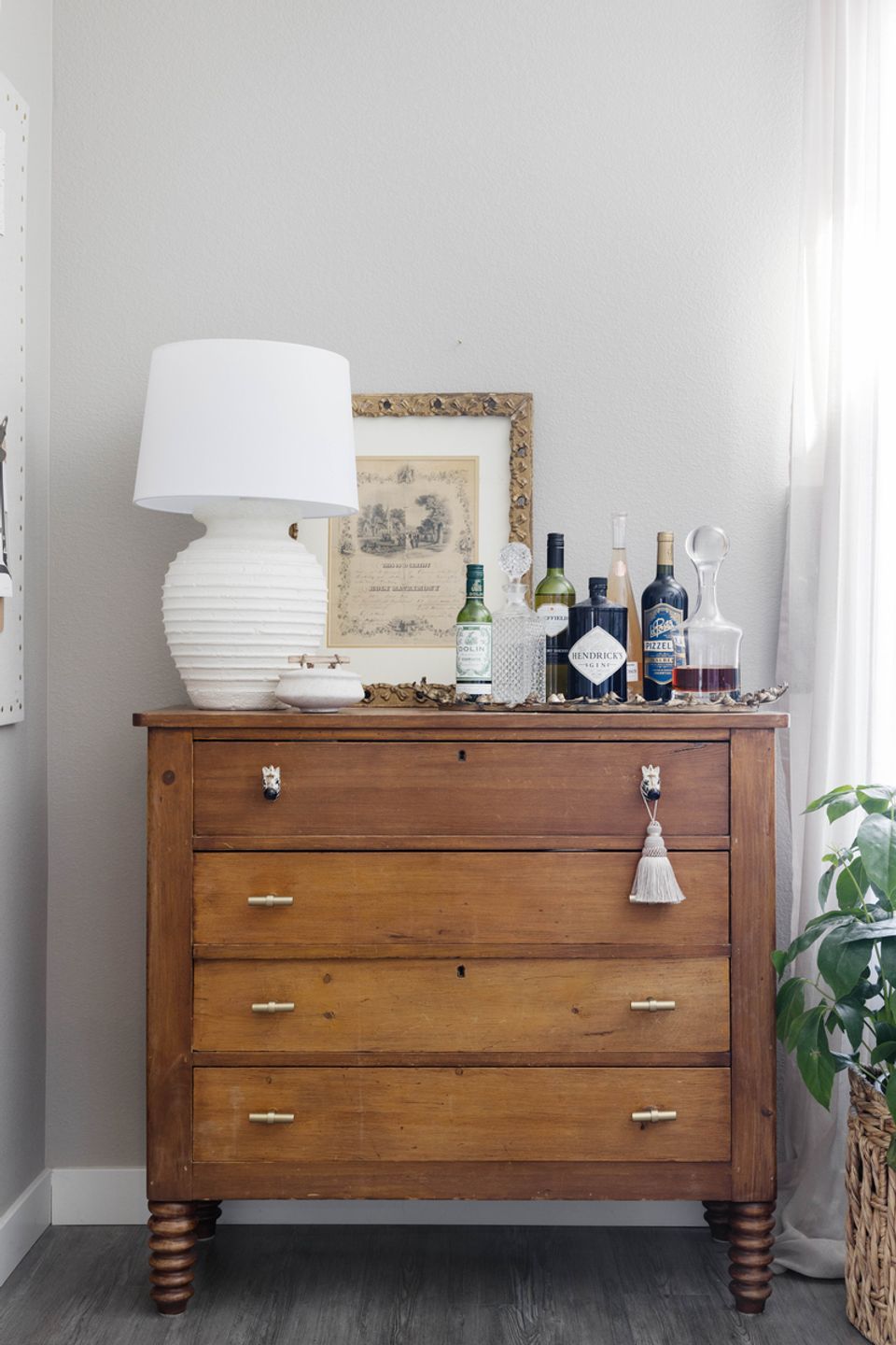 Warm wood dresser styled with white ceramic lamp, bottles, and vintage framed artwork against white walls