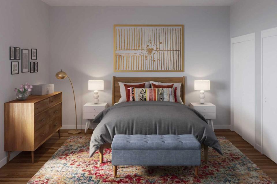 Warm mid-century bedroom with soft gray walls, natural wood furniture, layered bedding in charcoal and blue, and a colorful patterned rug creating cozy sophistication.