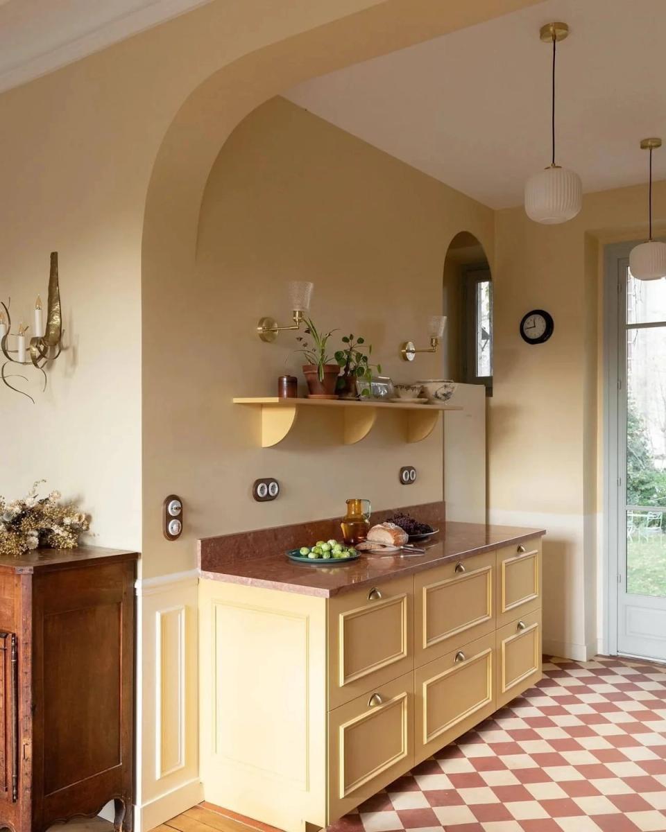 Butter-yellow transitional kitchen with arched niche, red-and-white checkerboard floor, and warm brass accents blending vintage elegance