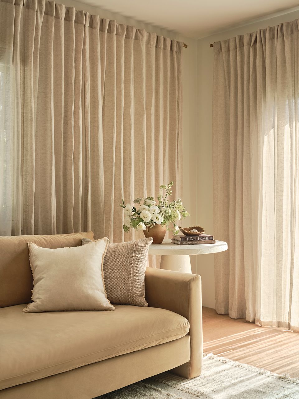 Serene neutral living room with curved beige sofa, layered cream and tan textiles, flowing floor-to-ceiling curtains, and minimalist botanical accents creating a calm, sophisticated atmosphere.