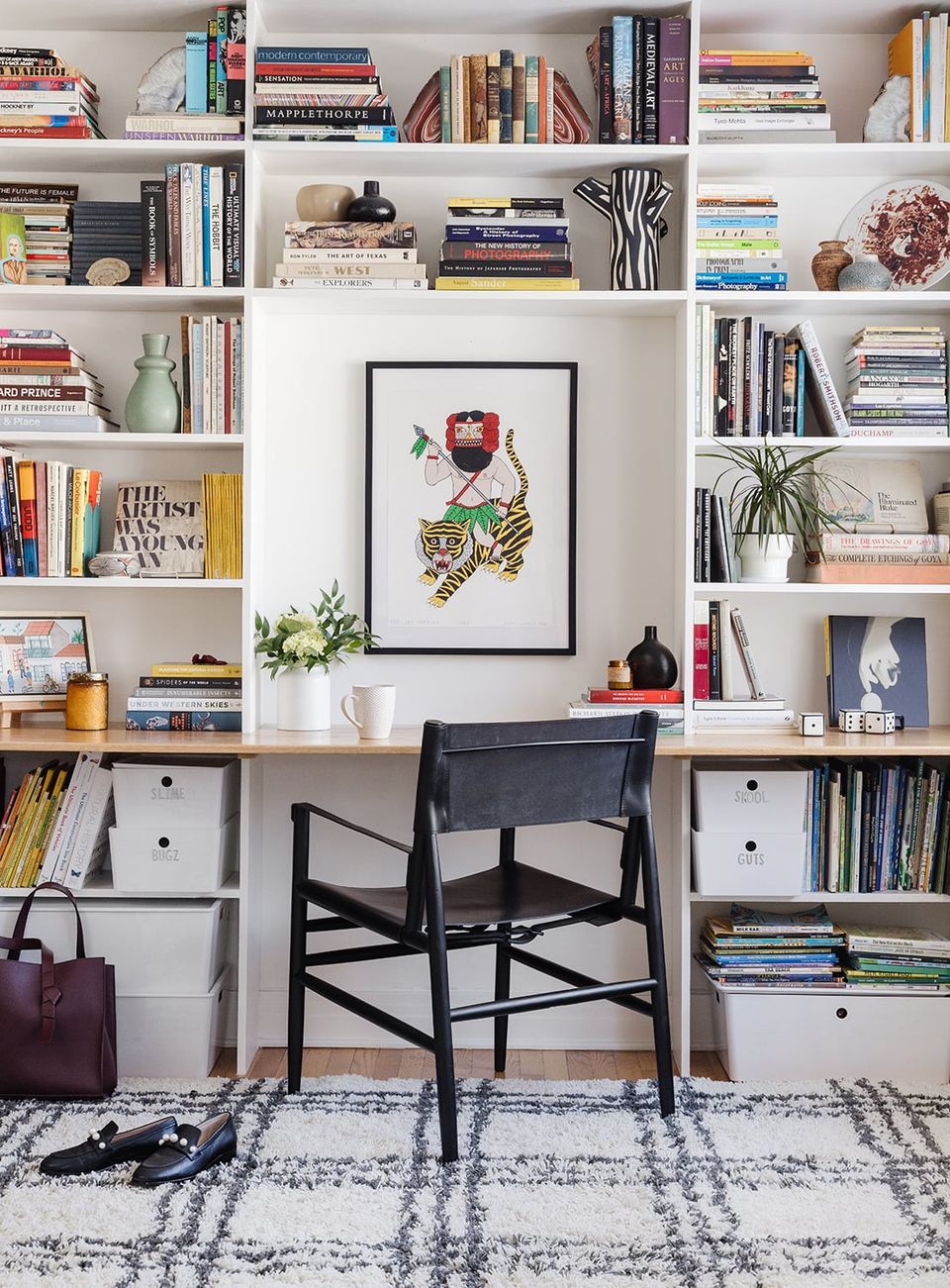 Contemporary minimalist workspace with white shelving, botanical artwork, black chair, patterned rug, and curated book collection