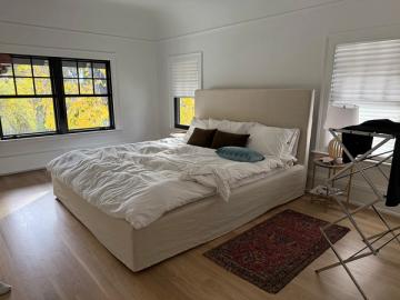 Minimalist modern bedroom with neutral upholstered bed, warm wood floors, and bright natural light from windows