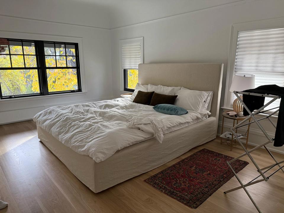 Minimalist modern bedroom with neutral upholstered bed, warm wood floors, and bright natural light from windows
