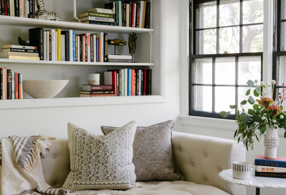 Bright library nook with white floating shelves, neutral upholstered window seat, layered throw pillows, and fresh greenery creating a serene reading retreat.