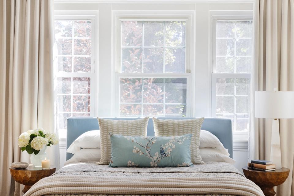 Sophisticated bedroom with slate blue headboard, layered neutral bedding, and abundant windows framing a serene, light-filled retreat.