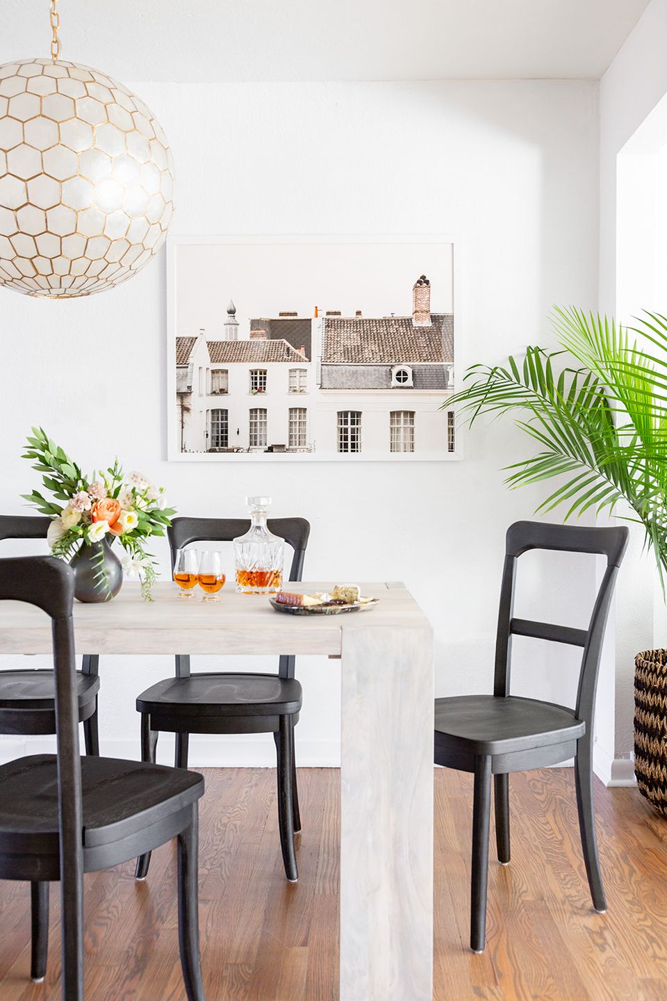 Bright Scandinavian dining space with white round table, black chairs, geometric pendant light, and tropical greenery accents