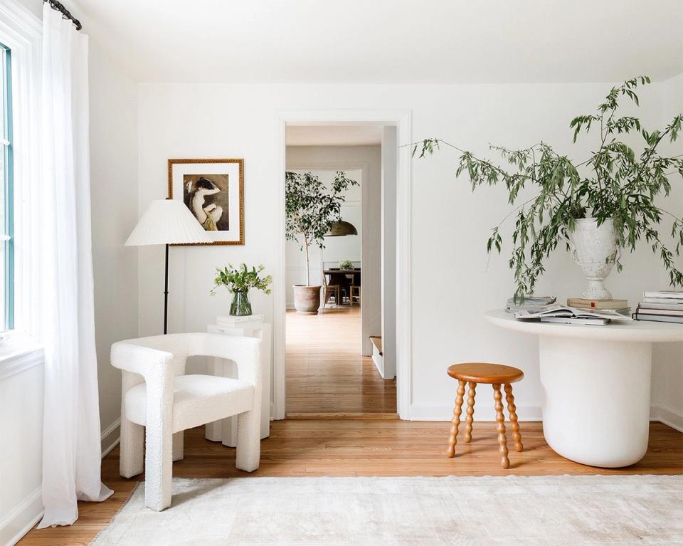 Minimalist modern living space with cream rounded chair, white cylindrical side table, and abundant greenery against soft neutral walls and natural light.