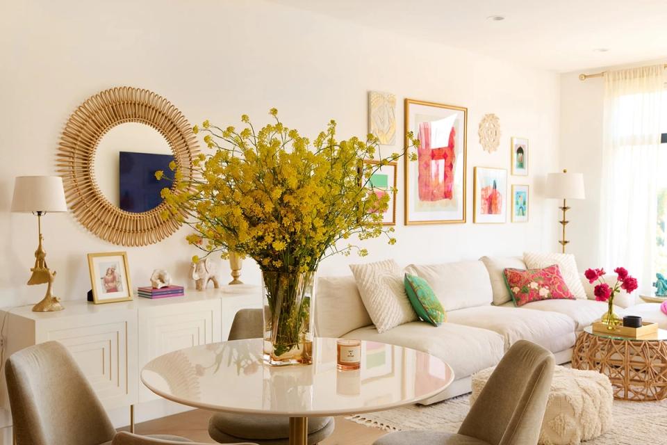 Cheerful living room with warm cream palette, vibrant yellow floral arrangement, woven mirror, and colorful artwork creating an inviting, eclectic mood