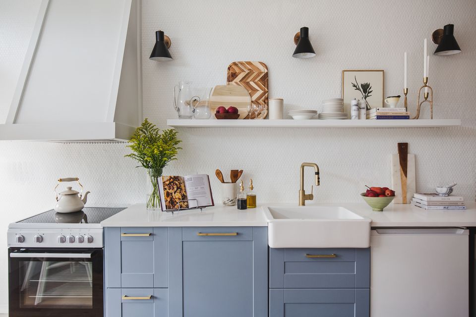 Bright minimalist kitchen with soft blue cabinetry, white subway tile, brass fixtures, and styled open shelving creating an airy, organized feel.