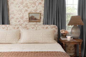 Traditional bedroom with floral toile wallpaper, cream patterned bedding, gray curtains, and warm wood nightstand creating classic elegance.