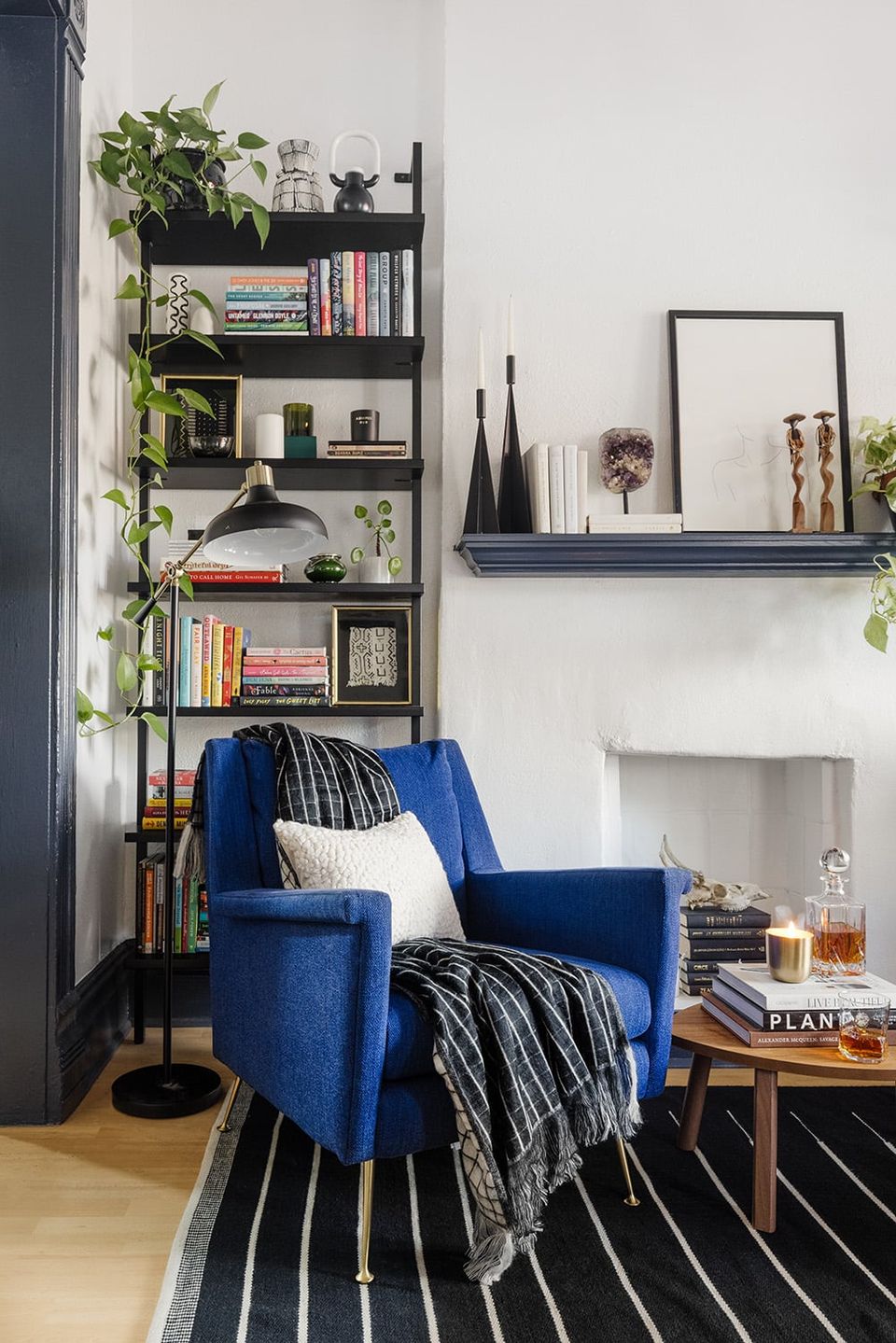 Modern reading nook with cobalt blue armchair, black metal shelving unit styled with plants and books, neutral walls, and striped area rug