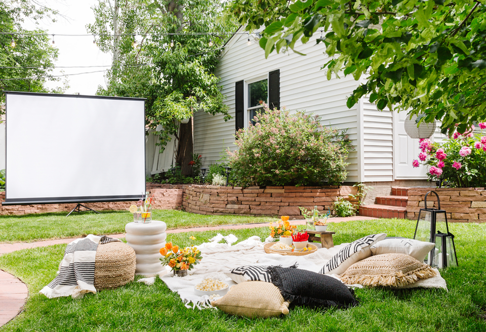 Backyard movie night setup with outdoor projector screen, cozy floor cushions, and garden flowers creating a casual, entertaining atmosphere