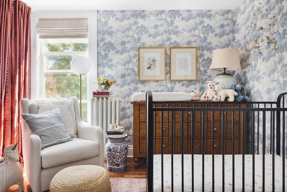 Nursery blending blue toile wallpaper, coral curtains, dark wood crib, and layered small-scale patterns in soft, sophisticated color palette