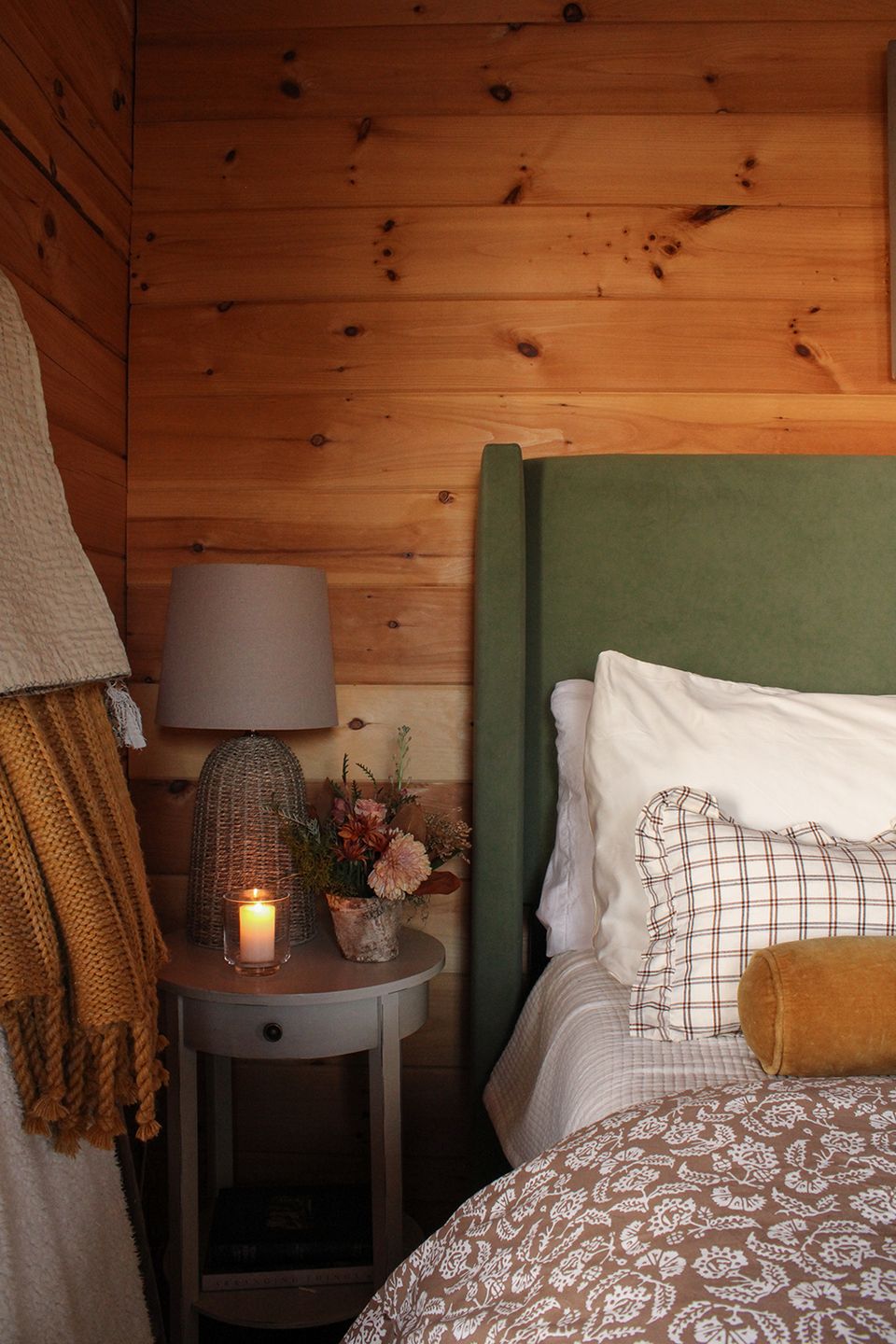 Rustic cabin bedroom: warm wood walls, olive green headboard, cozy layered textures, candlelit ambiance