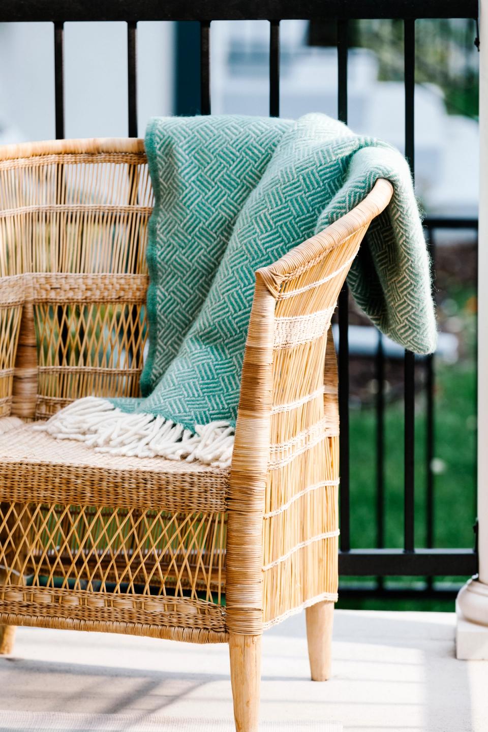 Coastal-inspired balcony chair with natural wicker frame, sage green geometric throw blanket, and layered textures creating a relaxed, inviting atmosphere.