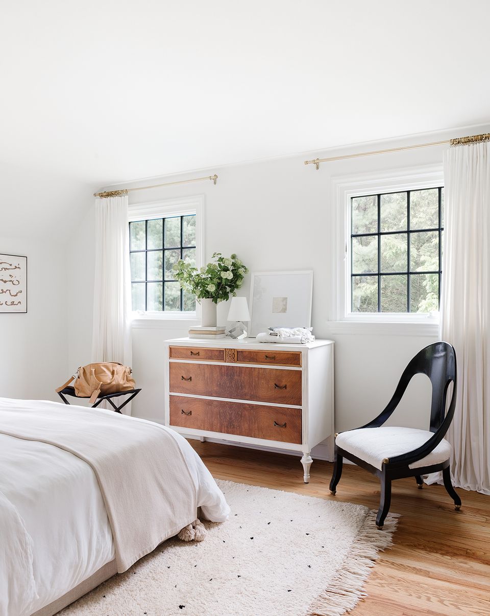 Serene French bedroom with white linens, wood dresser, black chair, and bright windows.