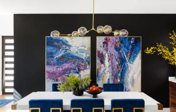 Bold dining space with navy stools, black wall backdrop, abstract colorful art, and gold pendant lighting for striking contrast