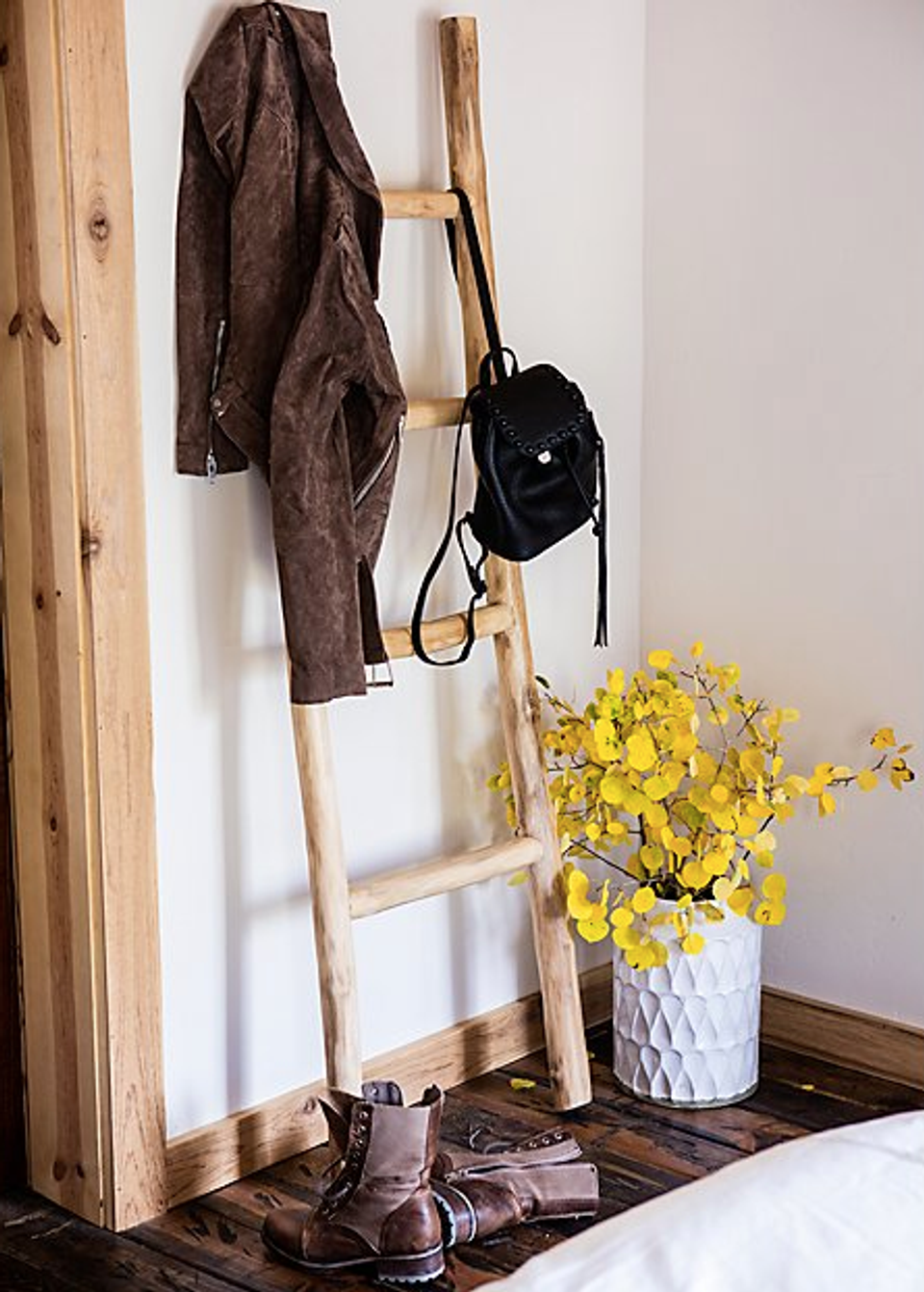 Rustic wooden ladder displaying brown jacket and black bag, styled with yellow flowers in textured white vase for functional farmhouse storage.