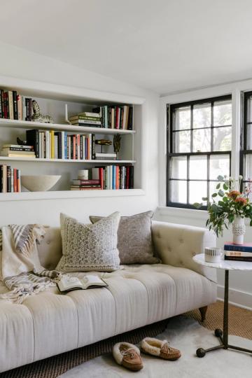 Minimalist library alcove with cream upholstered sofa, built-in white shelving filled with books, black-trimmed windows, and soft neutral palette
