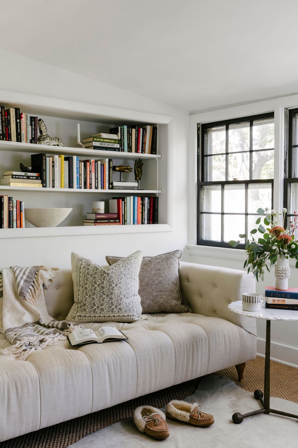 Minimalist library alcove with cream upholstered sofa, built-in white shelving filled with books, black-trimmed windows, and soft neutral palette