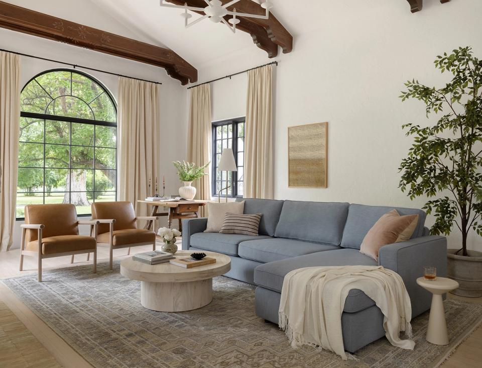 Transitional living room with exposed beams, blue sofa, caramel leather chairs, and warm neutral palette with greenery