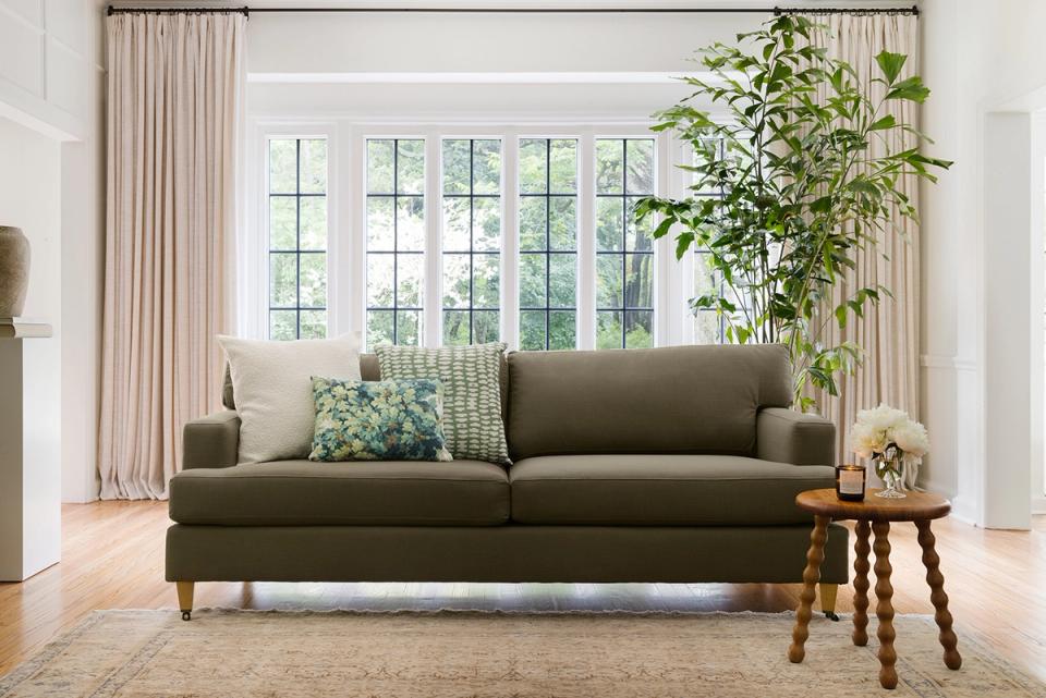 Contemporary living room with olive-green sofa, green geometric pillow accent, and lush potted plant against cream curtains and bright windows