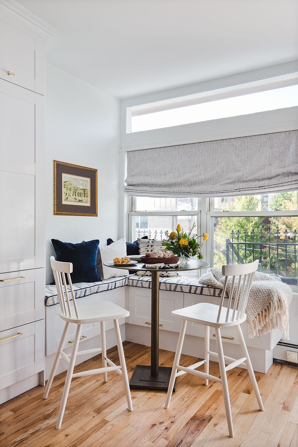 Minimalist window nook dining area with white spindle chairs, natural wood table, and navy blue accent pillow