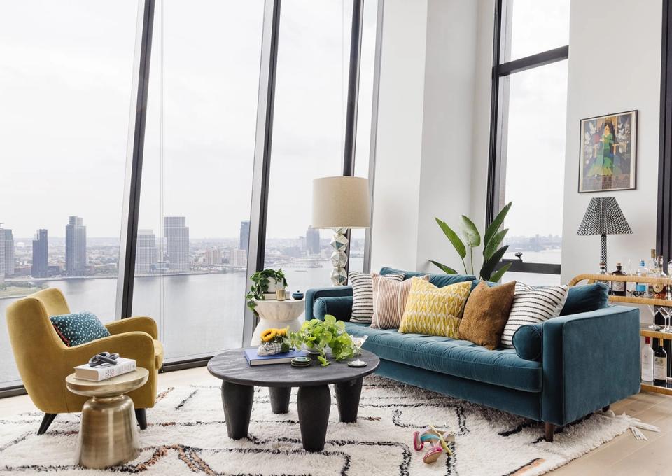 Modern minimalist living room with teal sectional, mustard accent chair, and city skyline views through floor-to-ceiling windows