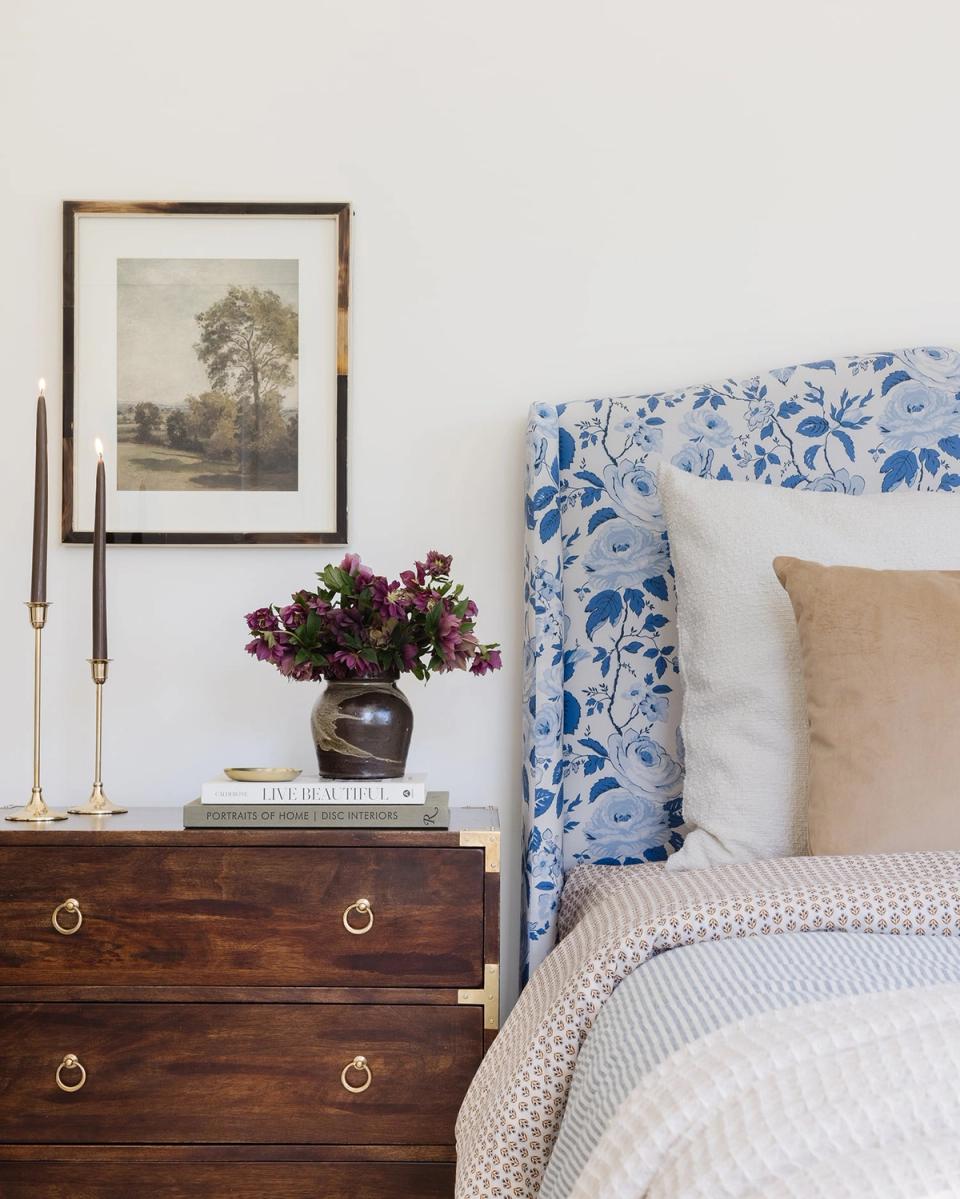 Classic bedroom style featuring blue and white patterned headboard, cream bedding, wood nightstand with brass hardware, and botanical artwork