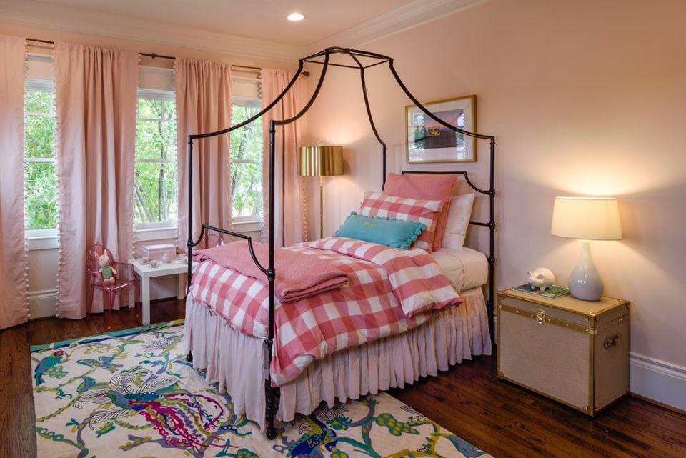 Whimsical girl's bedroom with wrought-iron canopy bed, red gingham bedding, soft pink walls, and colorful patterned rug.