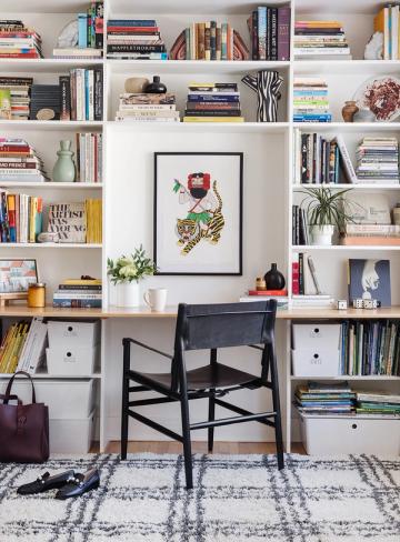 Gallery-style office with floor-to-ceiling white shelving, curated books, botanical art, and creative workspace