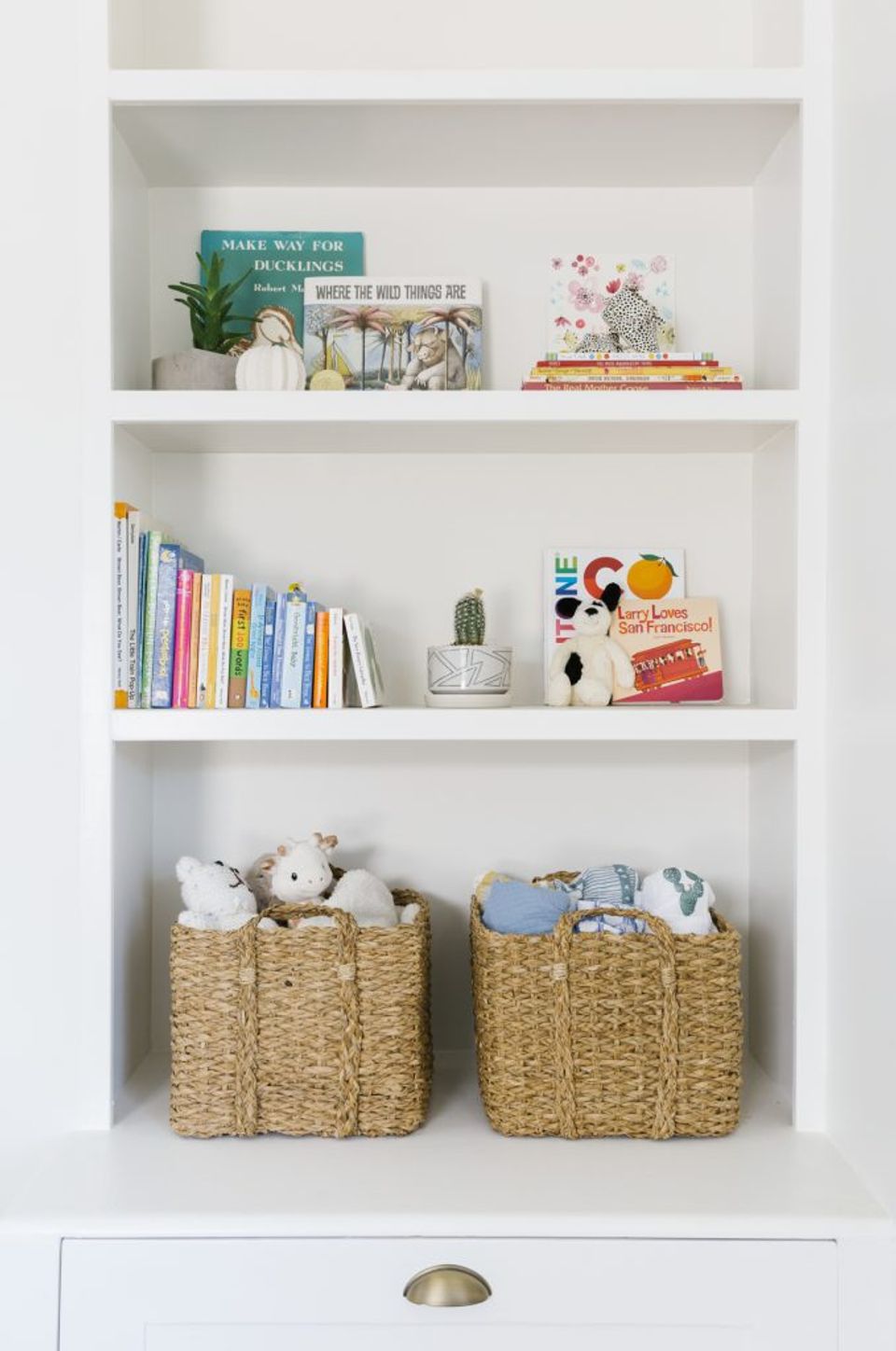 Open shelving with organized children's books, plants, and woven storage baskets in a bright, functional Scandinavian-inspired space.
