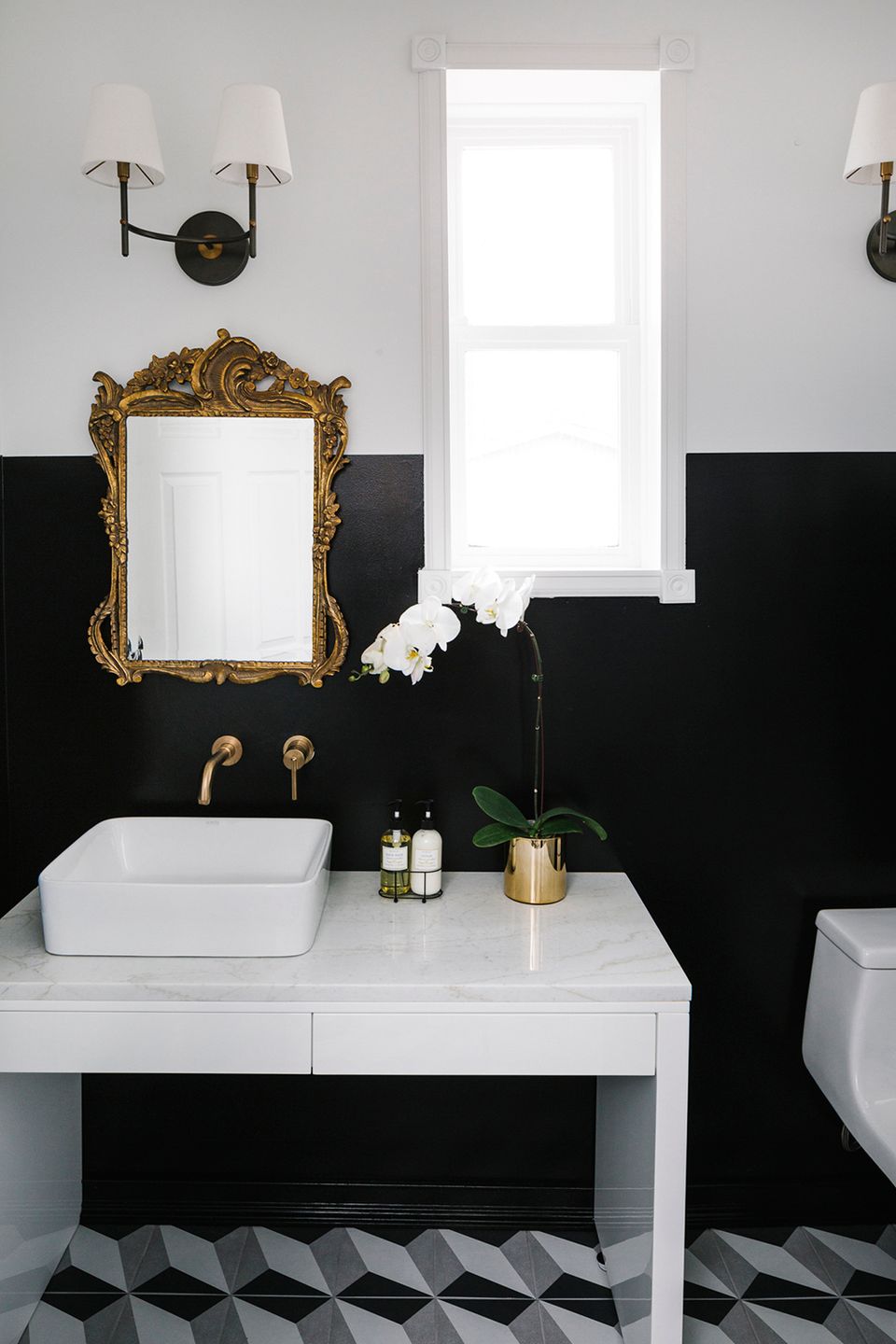 Bold, moody bathroom with dramatic black lower walls, white upper half, ornate gold mirror, and geometric tile floor for high-contrast glamour.