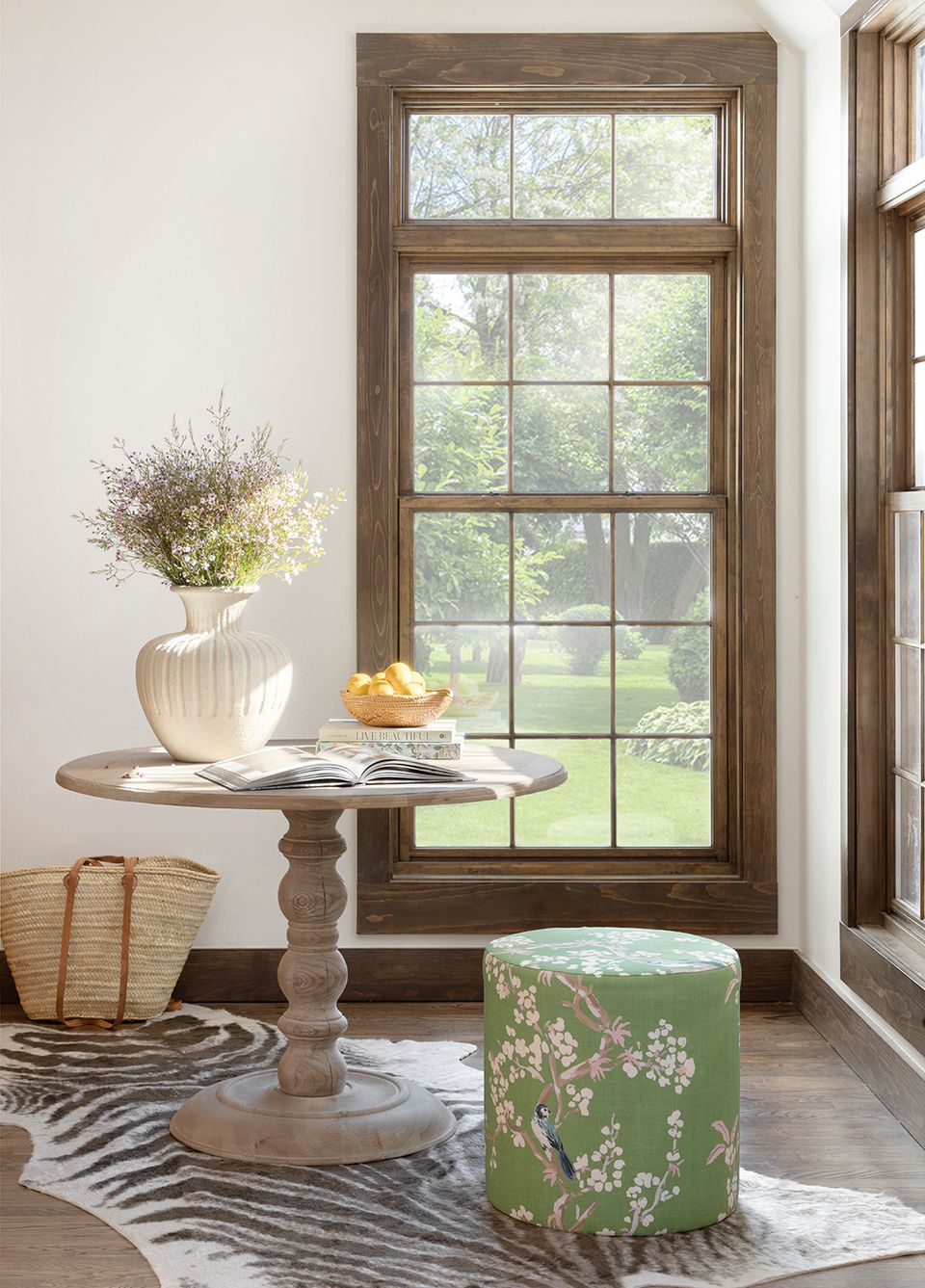 Farmhouse vignette with natural wood pedestal table, green floral ottoman, and neutral pottery against large multi-pane window