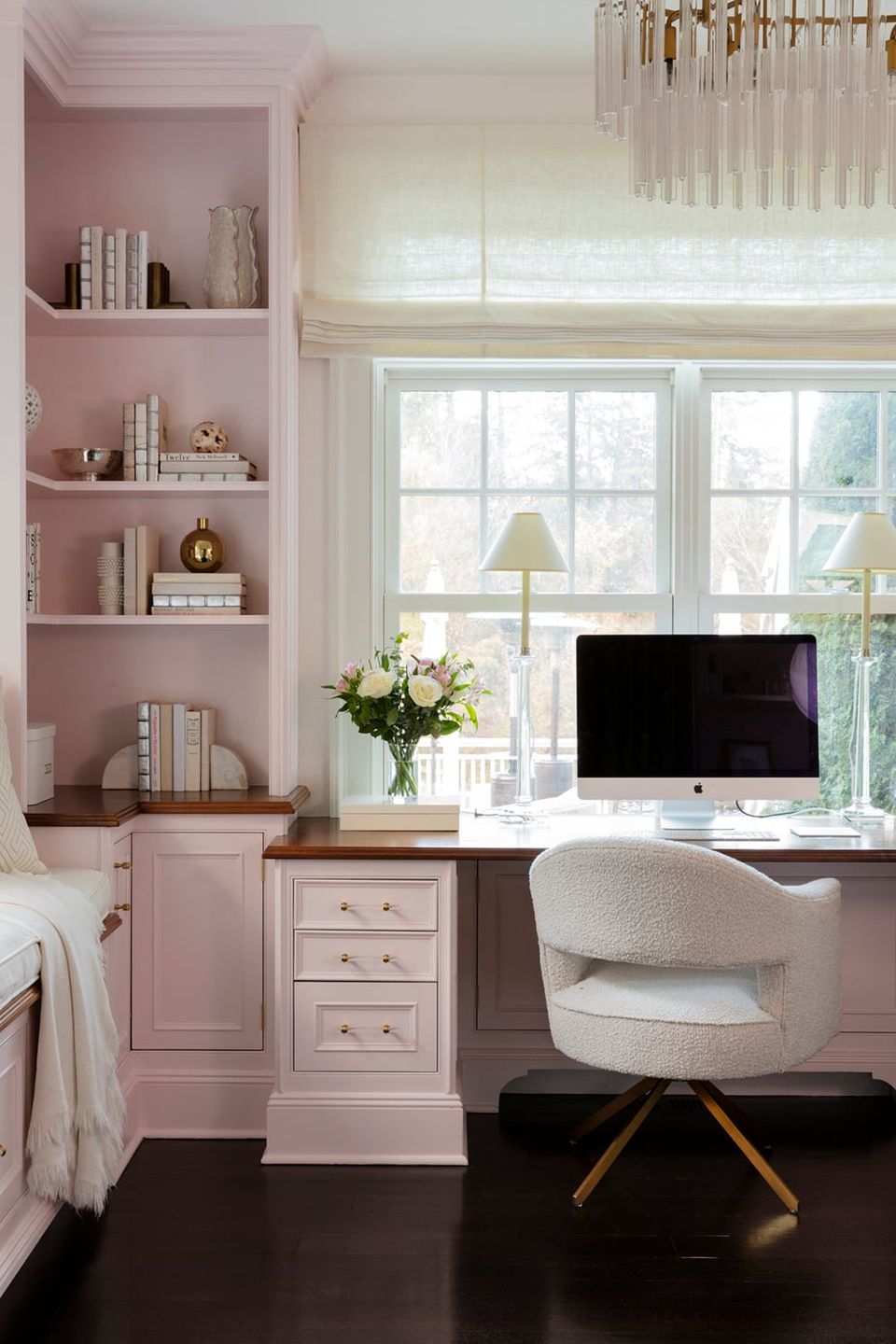 Feminine home office with soft pink built-in shelving, white marble desk, cream curved chair, and natural light from large windows