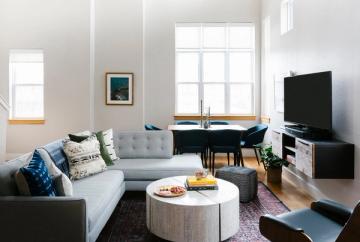 Contemporary minimalist living room combining TV area with home office, light gray upholstery, natural wood elements, and serene neutral color scheme