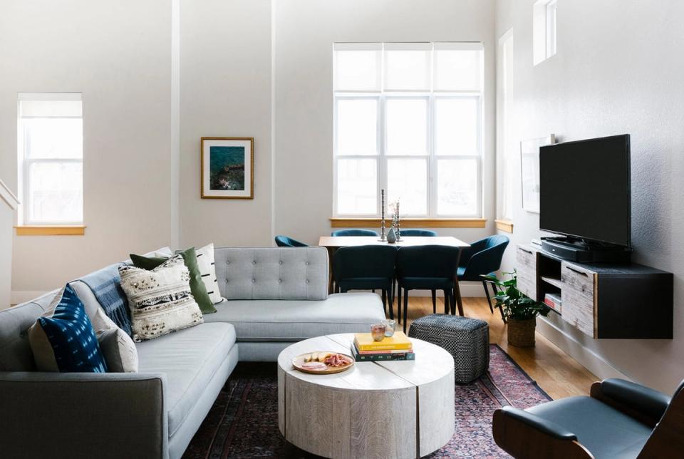 Contemporary minimalist living room combining TV area with home office, light gray upholstery, natural wood elements, and serene neutral color scheme