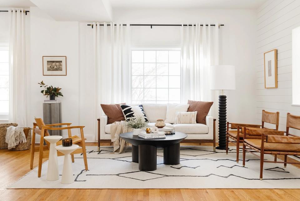 Bright eclectic modern living room with warm wood chairs, cream sofa, black coffee table, and curated art on white walls