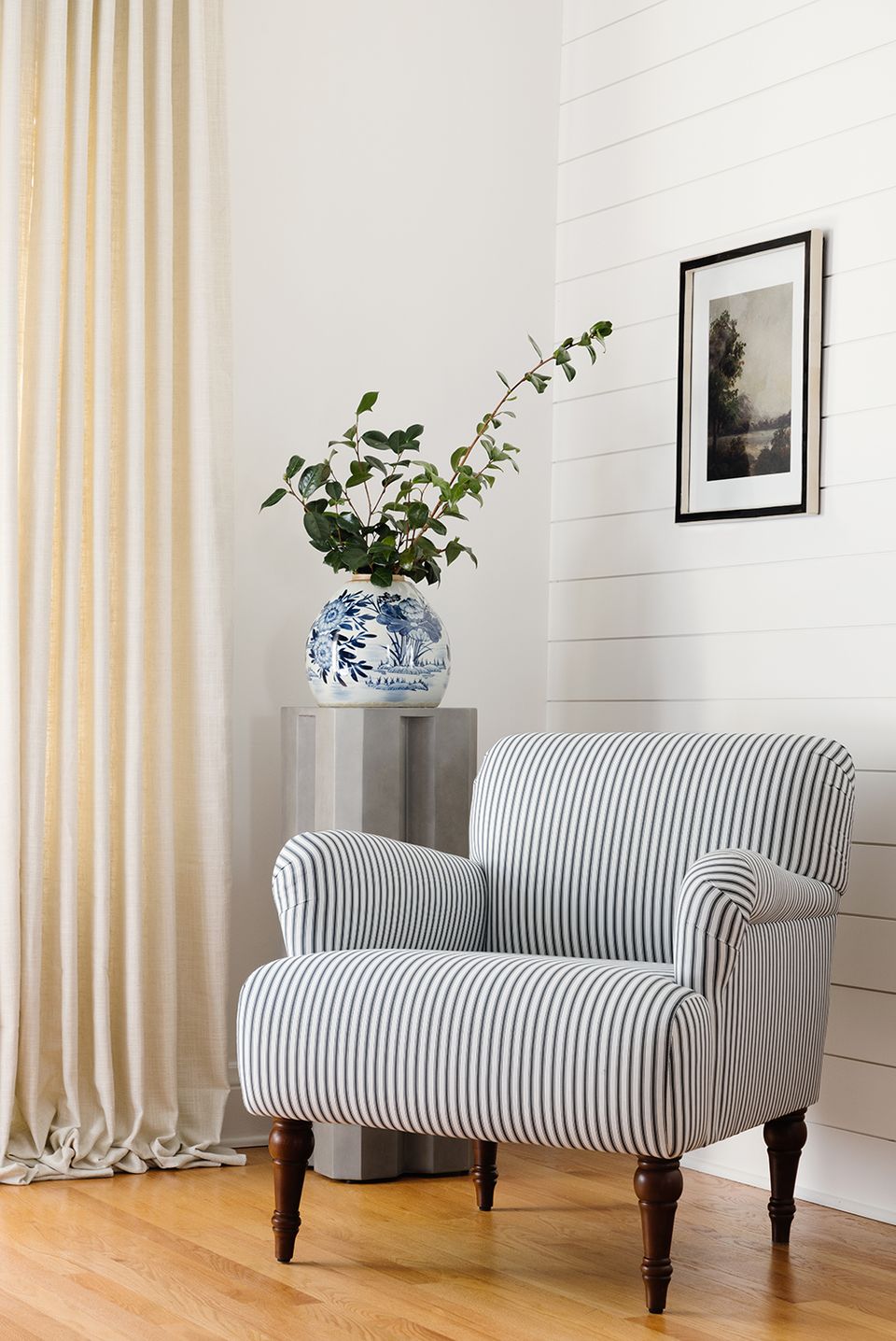 Farmhouse bedroom corner with striped accent chair, blue ceramic vase, black-framed landscape art, and cream curtains
